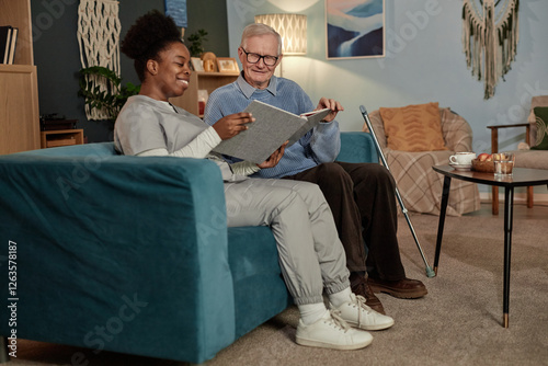 Full length of happy elderly man sitting on couch with female caregiver looking through vintage photograph album in living room during home care visit