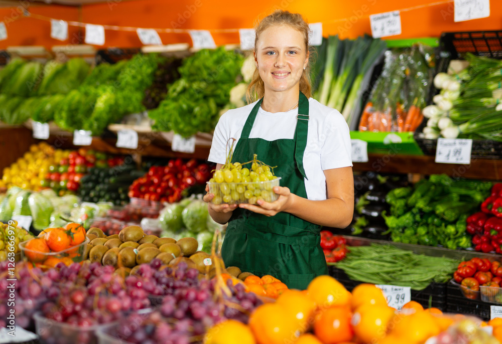 Portrait of a smiling fifteen-year-old girl who works part-time in a store as a trainee seller, standing at the counter, ..holding a box of grapes in her hands Portrait of a smiling fifteen-year-old girl who works part-time in a store as a trainee seller, standing at the counter, ..holding a box of grapes in her hands