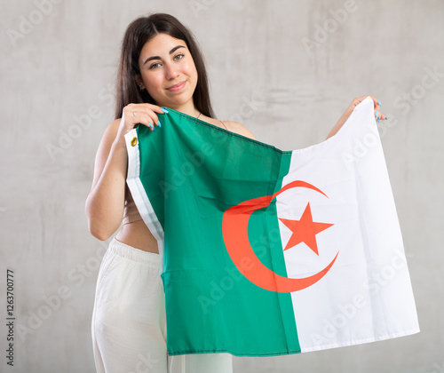 Happy young woman holding large flag of Algeria posing against background of light unicoloured wall