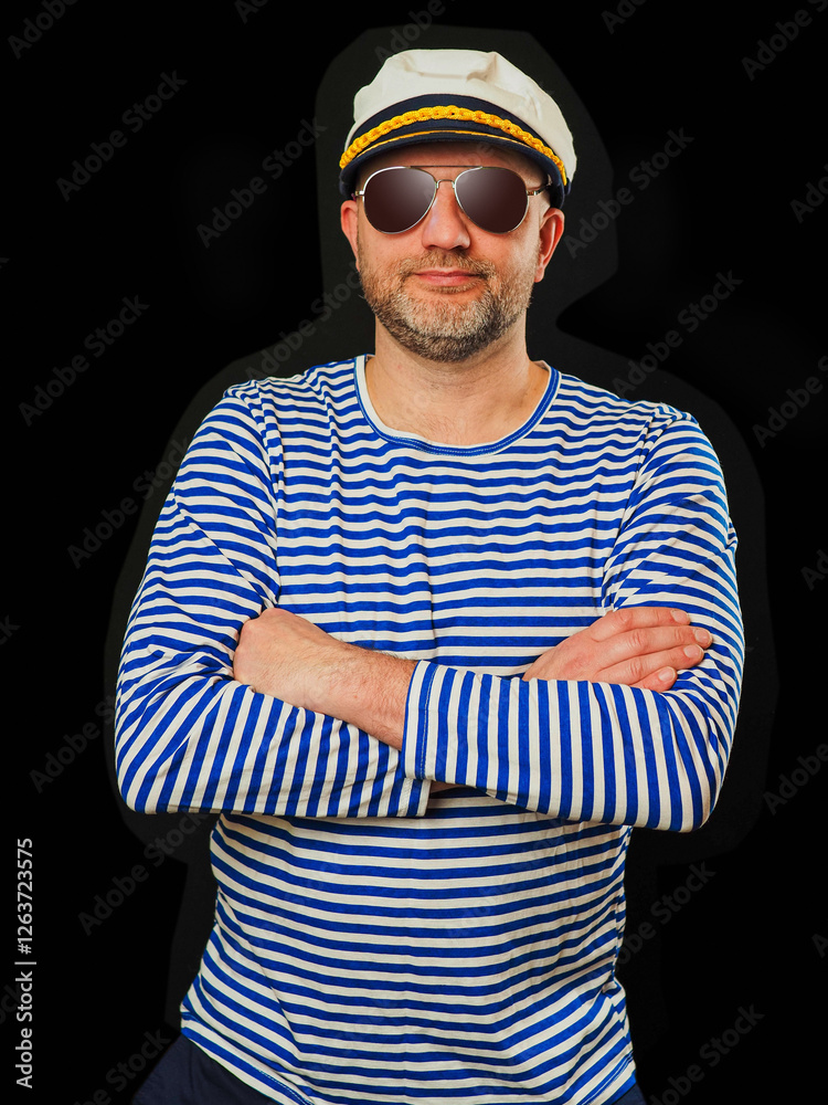 Fototapeta premium Portrait of a sailor in blue and white stripes shirt and captain hat. Male in his 40s with grey beard and dark glasses, hands crosses, smile on his face. Ocean theme party clothes.