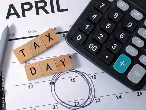 Overhead closeup top view photo of United States Tax Day calendar with circled 15th date, TAX DAY text letters in wood tiles. Black accounting calculator with white numbers.
