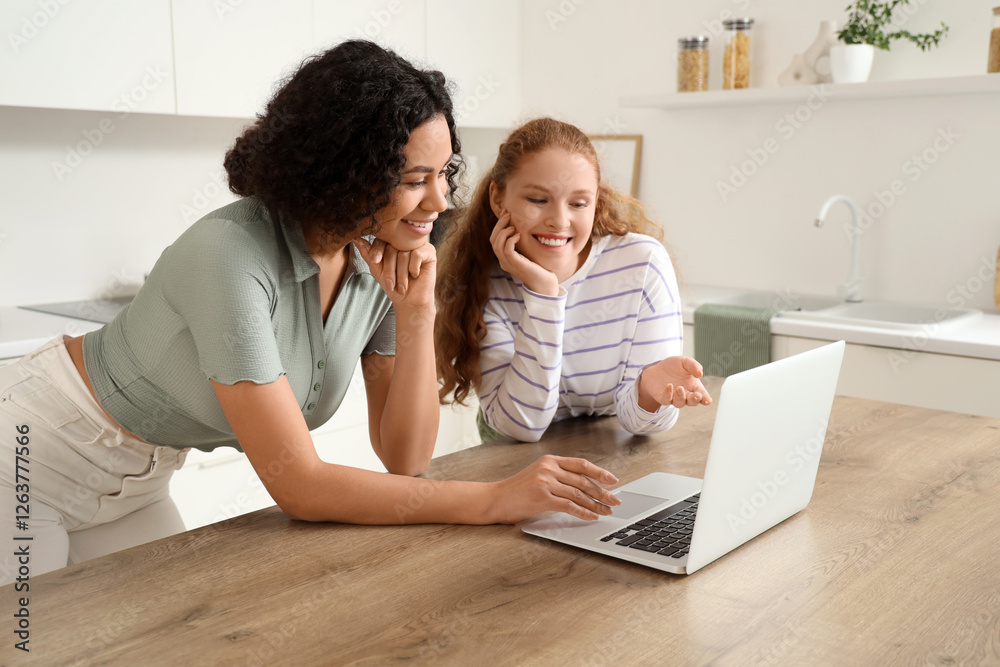 Beautiful young happy female friends with laptop sitting in kitchen at home Beautiful young happy female friends with laptop sitting in kitchen at home