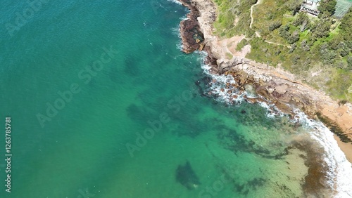 Drone shot aerial view of clear blue water and waves crashing onto rocks near a beach in summer in Australia 