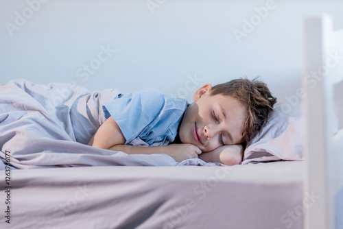 A young boy wearing a blue shirt is peacefully sleeping on his cozy bed tucked in with soft blankets and surrounded by comfortable linens.