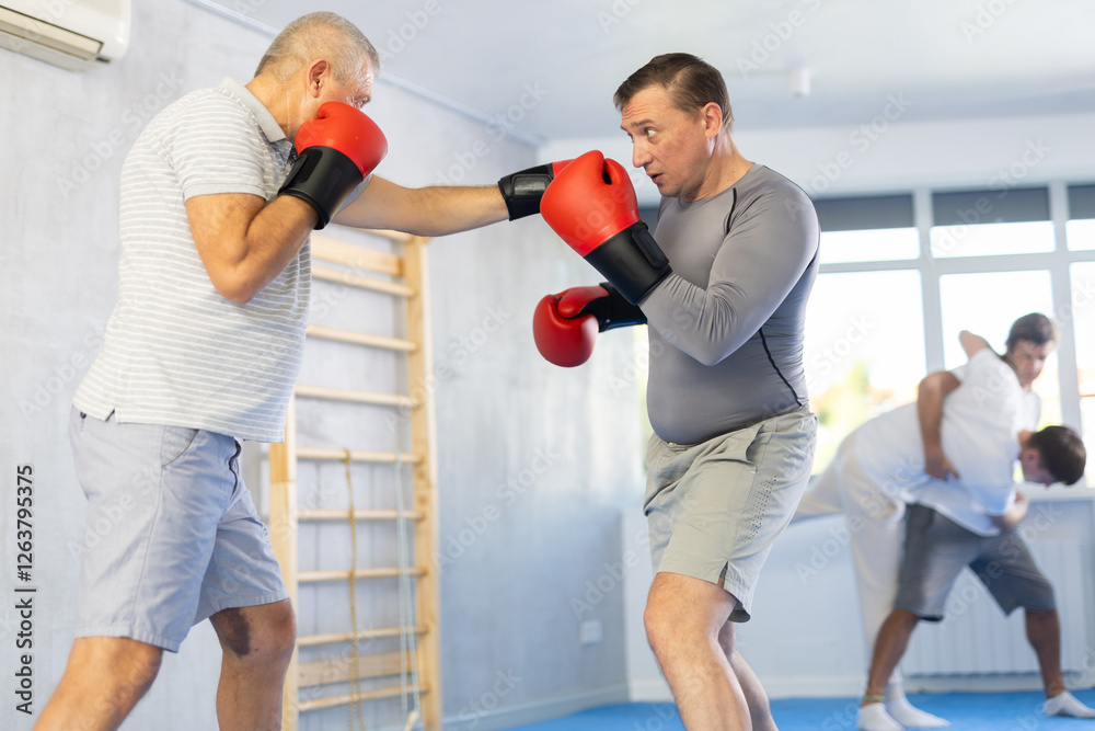 Mature man and male partner in boxing gloves punch deliver blow each other during training sparring session. Practical use of technique and force of impact in combat practice. Mature man and male partner in boxing gloves punch deliver blow each other during training sparring session. Practical use of technique and force of impact in combat practice.