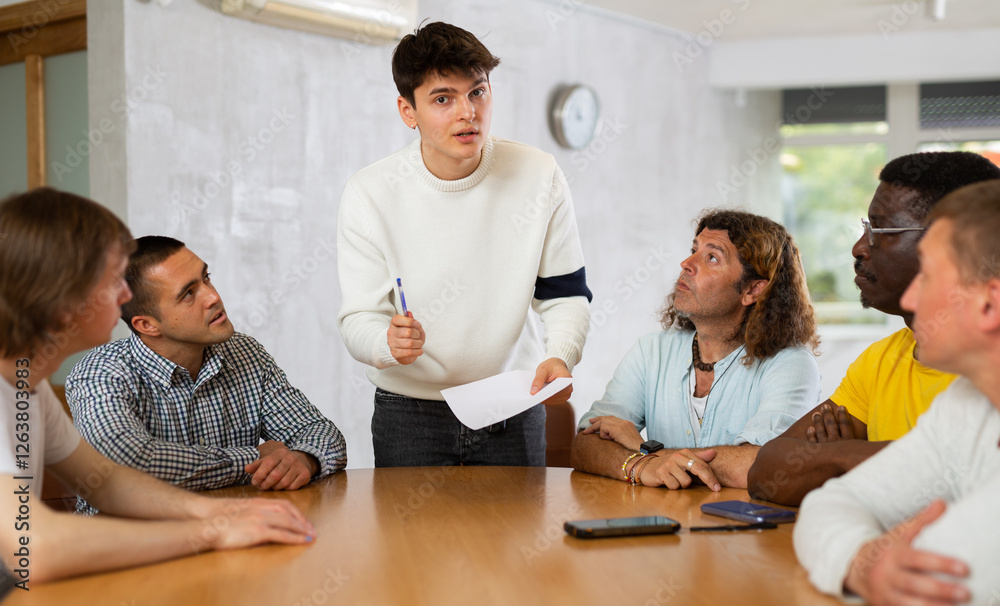 Focused young guy student holding papers ad discussing assignments or projects with interested male group of various ages and nationalities gathered around table in classroom.. Focused young guy student holding papers ad discussing assignments or projects with interested male group of various ages and nationalities gathered around table in classroom..