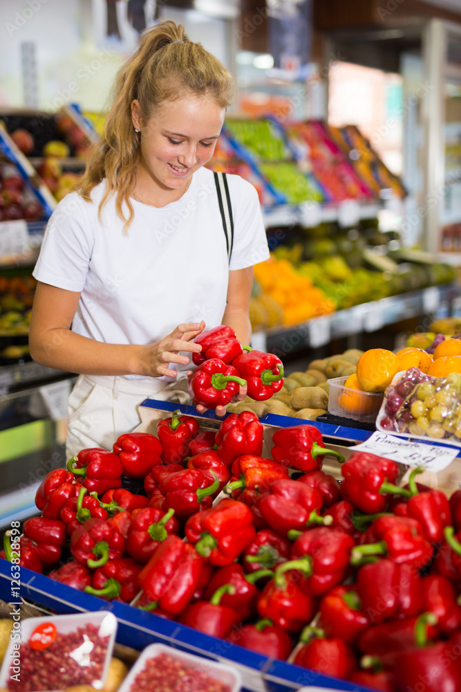 Fifteen-year-old girl who came to the supermarket for shopping, chooses bell pepper, standing near the counter Fifteen-year-old girl who came to the supermarket for shopping, chooses bell pepper, standing near the counter