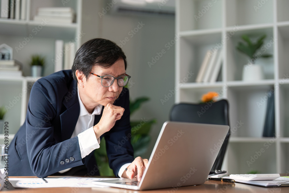 Elderly businessman deep in thought while working on a laptop in a modern office. Elderly businessman deep in thought while working on a laptop in a modern office.