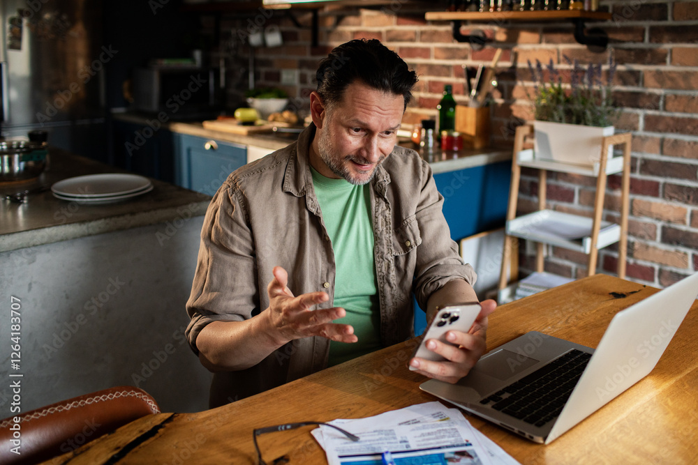 Man working from home talking on smartphone in kitchen with laptop and documents Man working from home talking on smartphone in kitchen with laptop and documents