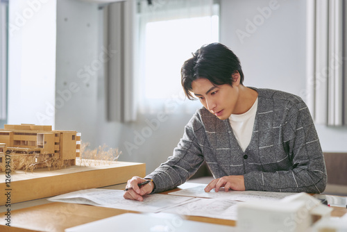 建築の模型を横に都市計画の為の設計図を書く工学部の大学の建築学科で勉強する大学生の男性