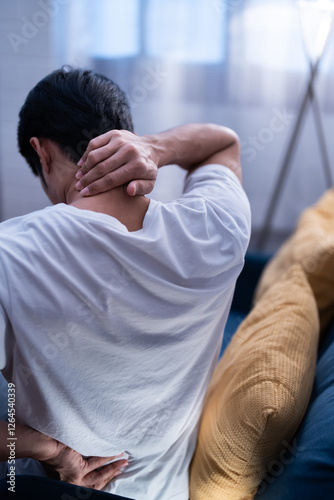 Asian freelancer man sitting on a couch holding his neck and lower back in discomfort, possibly due to muscle strain, poor posture, or stress and tired of working