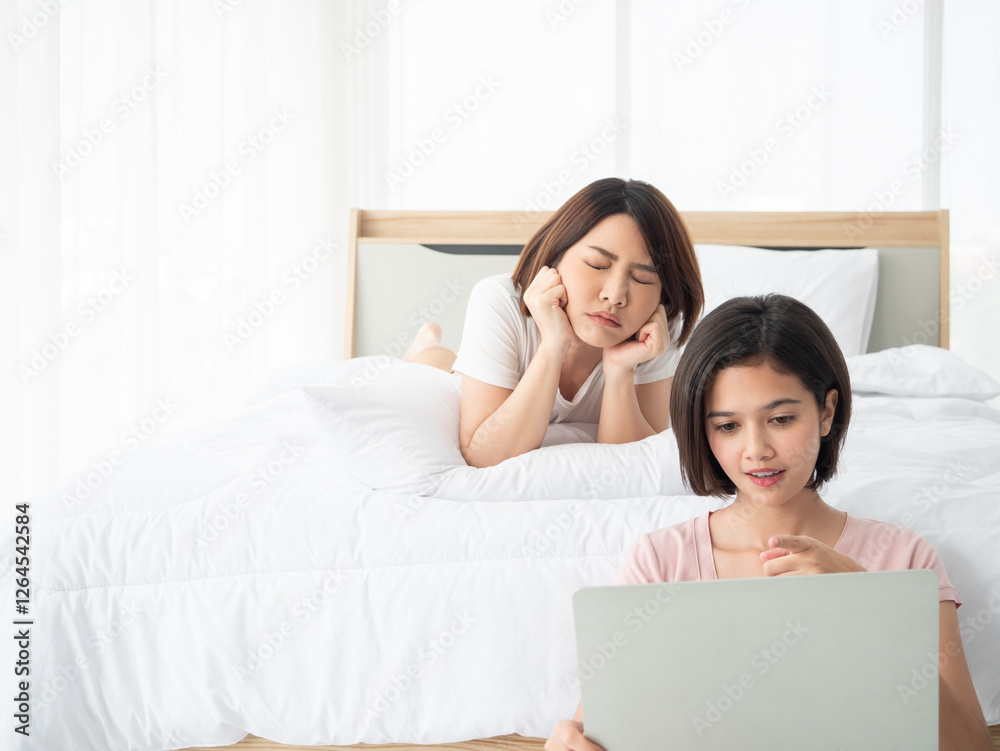 Asian young woman looks bored while lying on a bed, resting her face on her hands, as her friend focuses on a laptop Asian young woman looks bored while lying on a bed, resting her face on her hands, as her friend focuses on a laptop