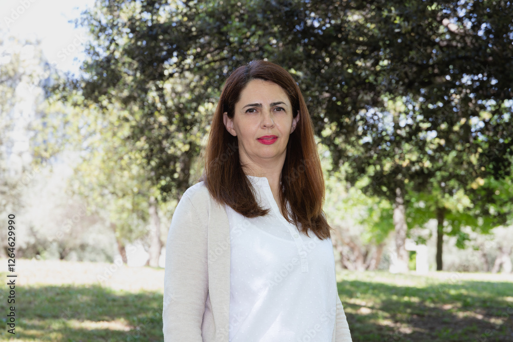 Portrait of mature woman looking at camera in a park with natural light. Serene expression, casual dress in light tones. Concept of well-being, nature, tranquility and healthy life Portrait of mature woman looking at camera in a park with natural light. Serene expression, casual dress in light tones. Concept of well-being, nature, tranquility and healthy life