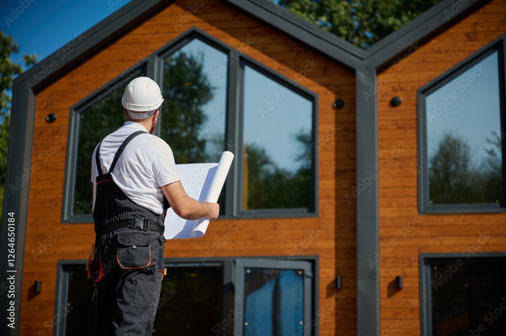 Architect hold plan. Bearded architect wearing hardhat holding project blueprints paper plans standing near comlex of new modern houses looking to something at construction site. Architect hold plan. Bearded architect wearing hardhat holding project blueprints paper plans standing near comlex of new modern houses looking to something at construction site.