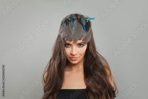 Glam female model with healthy long wavy hairstyle, trendy make-up and hairdeco feathers accessories on her head looking at camera, fashion beauty portrait photo