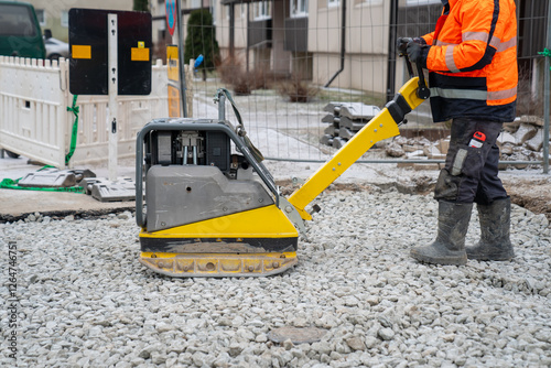 Yellow construction vibratory plate compactor on gravel for ground leveling job site project. Yellow construction equipment. Construction worker wearing vest and working outdoors and cold