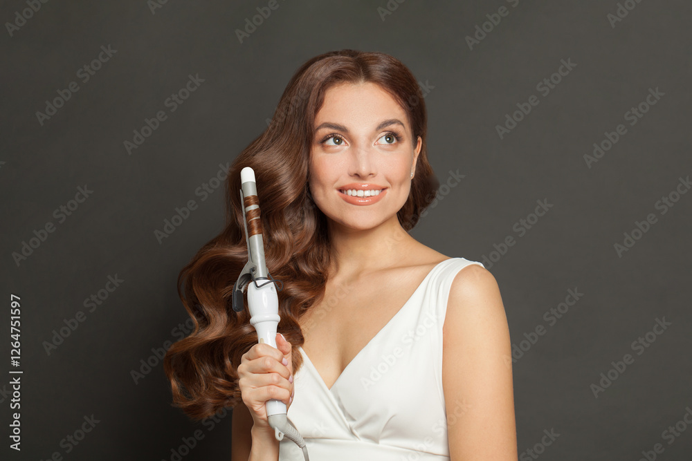 Cheerful brown haired woman with shiny long curly hairstyle showing curling iron. Hair treatment, hair care and styling Cheerful brown haired woman with shiny long curly hairstyle showing curling iron. Hair treatment, hair care and styling