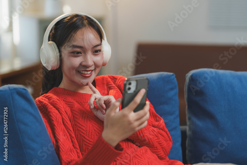 Smiling young woman wearing headphones, relaxing on a blue couch while browsing her smartphone. Her casual attire and joyful expression suggest leisure and entertainment