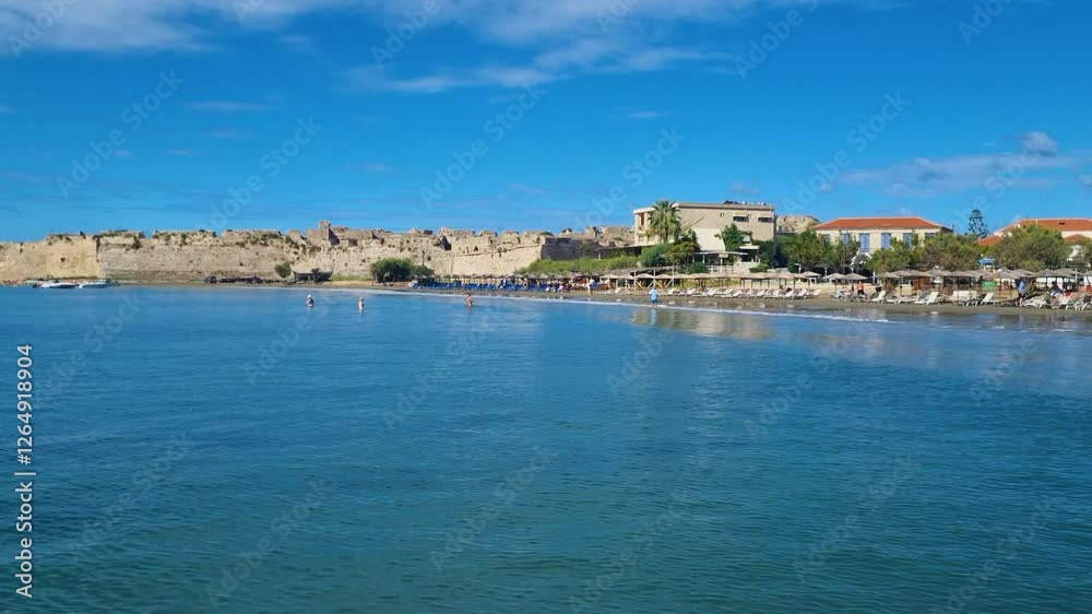 View from the sea towards the Venetian castle, the beach, and the town of Methoni on a beautiful, sunny summer day. In the distance, people enjoy bathing in crystal-clear waters of the Mediterranean View from the sea towards the Venetian castle, the beach, and the town of Methoni on a beautiful, sunny summer day. In the distance, people enjoy bathing in crystal-clear waters of the Mediterranean