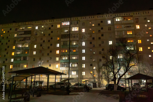 A large Soviet-style apartment building stands illuminated against the dark night sky, with its windows glowing in various warm and cool tones. The playground and parked cars are in the foreground 