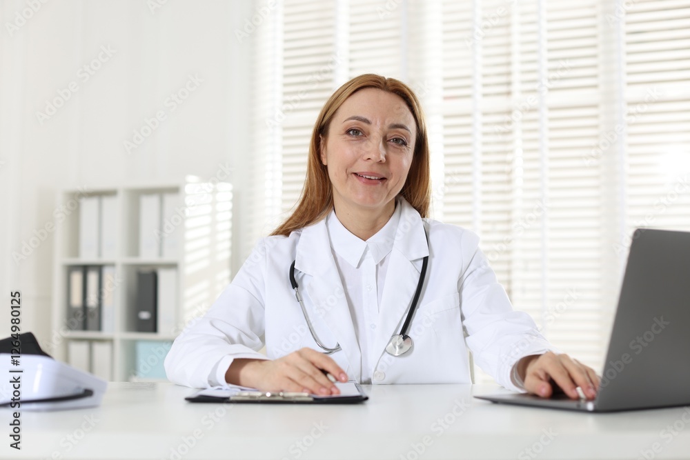 Cardiologist with stethoscope using laptop at white table in clinic Cardiologist with stethoscope using laptop at white table in clinic