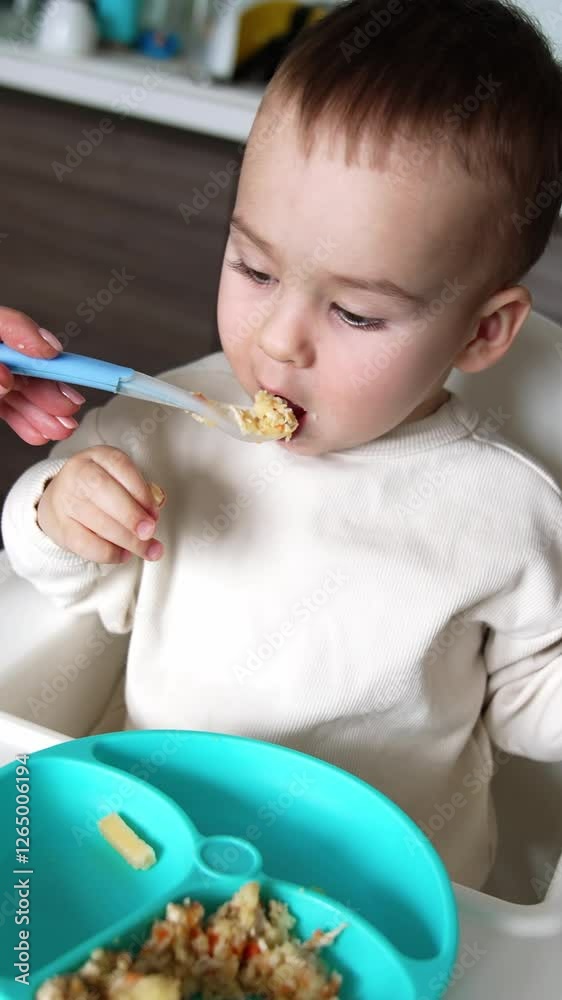 Mom shoves a full spoon to her son's mouth. Adorable kid takes a spoon from a table. Close up. Kitchen backdrop. Vertical video Mom shoves a full spoon to her son's mouth. Adorable kid takes a spoon from a table. Close up. Kitchen backdrop. Vertical video