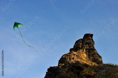 A kite in the blue sky, hovering next to a rock.