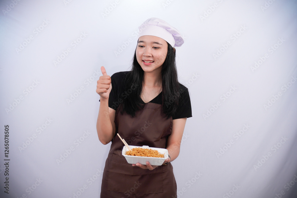 Asian woman wearing chef's uniform holding container of instant noodles with thumbs up sign Asian woman wearing chef's uniform holding container of instant noodles with thumbs up sign