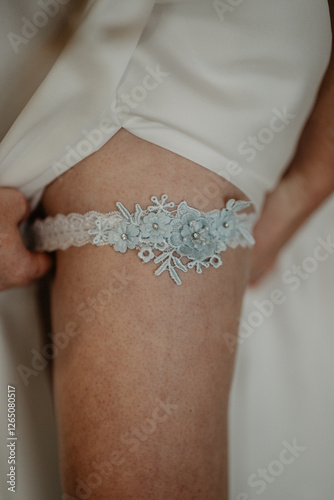  A close-up of a bride lifting her white wedding dress to reveal a delicate lace bridal garter adorned with intricate blue floral embroidery and sparkling rhinestones, symbolizing a timeless wedding 
