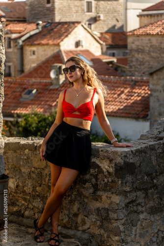 A stylish woman poses against the historic stone walls of a European old town. The warm sunlight highlights the rustic rooftops, creating a travel and fashion-inspired scene.