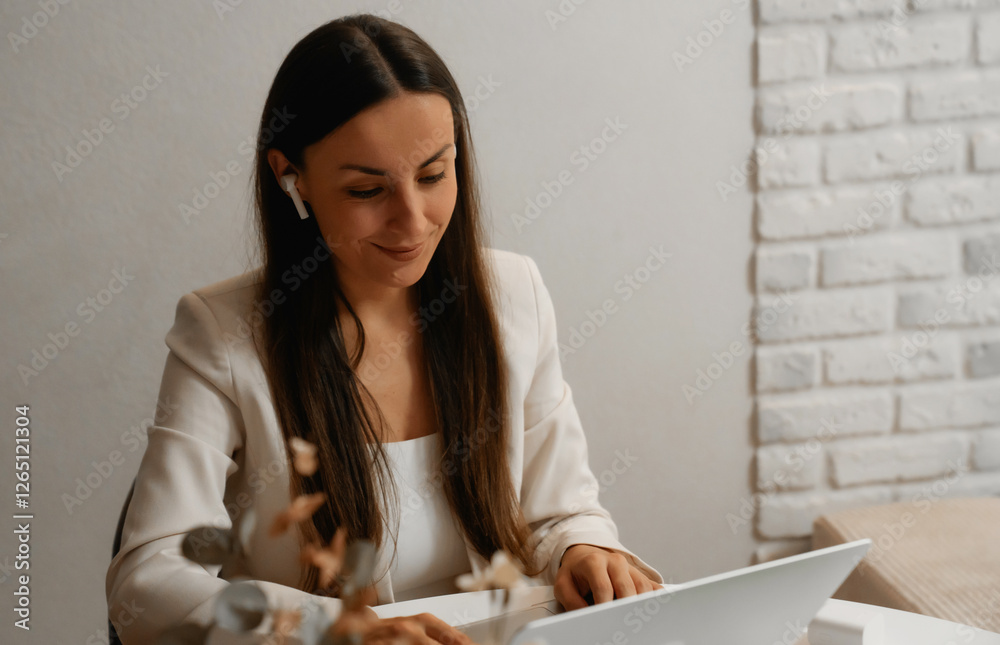 A woman in a white blazer smiles while working on her laptop, wearing wireless earbuds. She is focused, seated at a clean workspace with a minimalist background and a modern atmosphere. A woman in a white blazer smiles while working on her laptop, wearing wireless earbuds. She is focused, seated at a clean workspace with a minimalist background and a modern atmosphere.