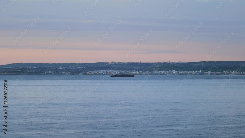 Stunning view of the Malta coastline at sunrise, featuring calm Mediterranean waters and a cargo ship in the distance. The soft morning light enhances the urban landscape and rolling hills. Stunning view of the Malta coastline at sunrise, featuring calm Mediterranean waters and a cargo ship in the distance. The soft morning light enhances the urban landscape and rolling hills.