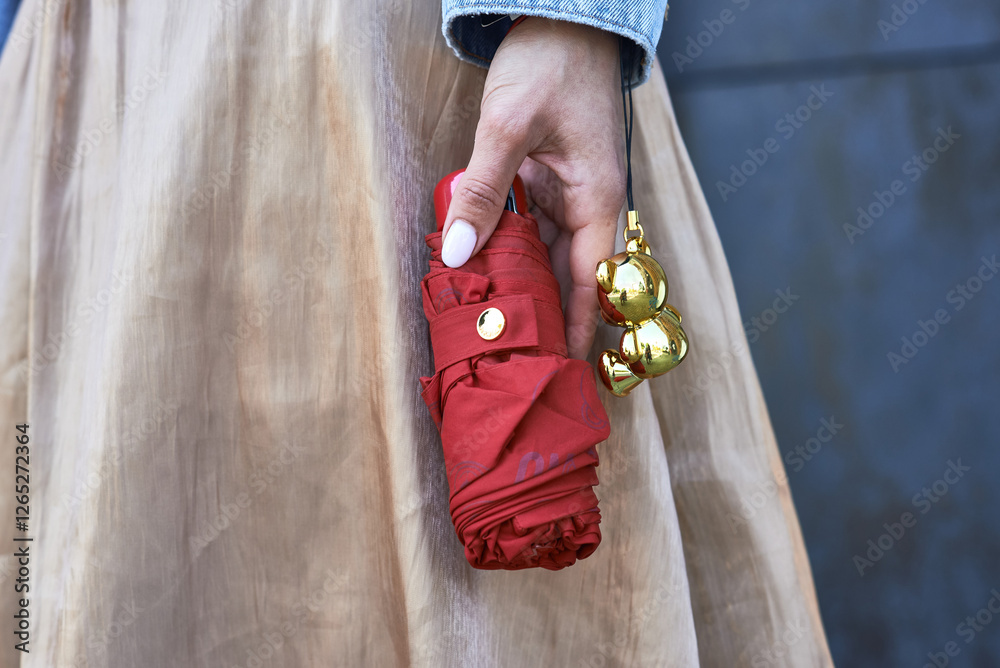 Hand holding red umbrella with golden ornament strapped on wrist against beige fabric backdrop, rainy day essentials concept