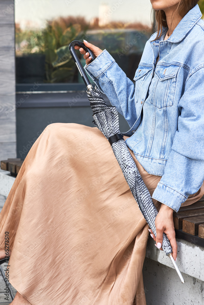 Woman in casual denim jacket and skirt holding patterned closed umbrella on bench, style concept