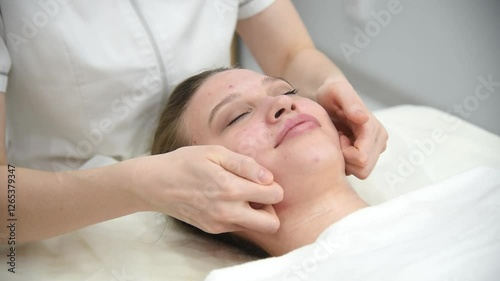 face massage, young attractive caucasian woman, beautiful relaxed lady with problem skin and red inflammations, white towel, spa therapist doing facial massage in beauty salon, skincare concept photo