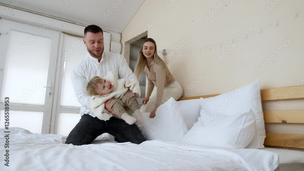Loving Caucasian father waves his laughing baby boy on the bed. Dad throws his kid into the pillows. Mother at backdrop smiles.