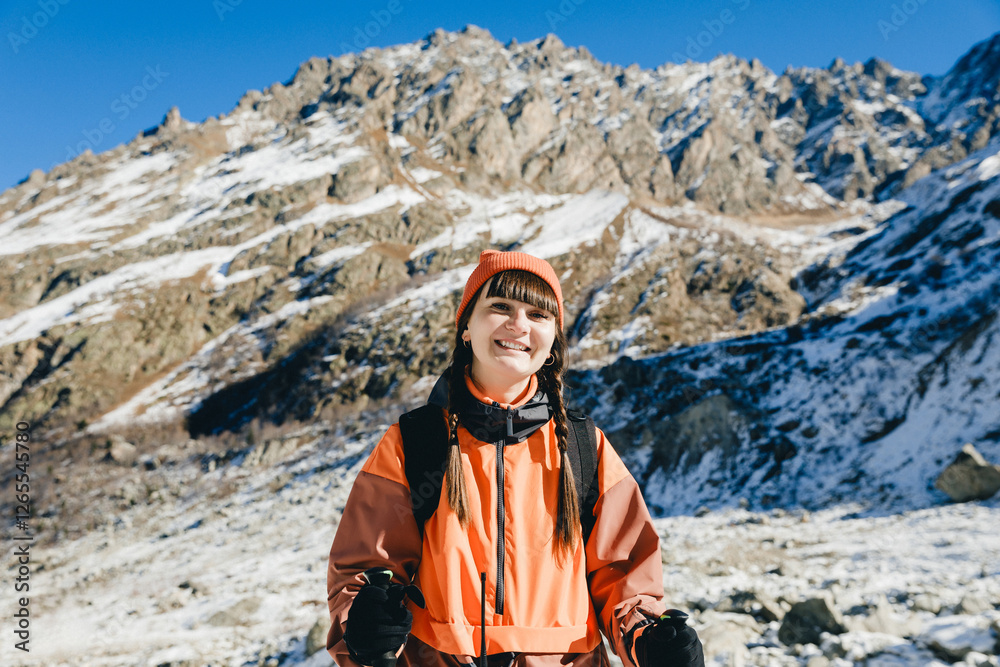 young woman in a winter hat, orange waterproof jacket and backpack standing against the backdrop of a winter mountain valley - adventure concept. woman trekking guide young woman in a winter hat, orange waterproof jacket and backpack standing against the backdrop of a winter mountain valley - adventure concept. woman trekking guide