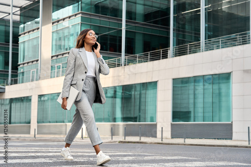 Full body portrait of successful African American busy business woman having phone call outdoors. Middle age businesswoman in suit talking on smartphone, walking near office centre. Banner, copy space