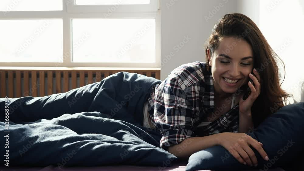 Young woman stay at home during quarantine. Typing phone number on smartphone screen and talking with pleasure. Lying on bed in morning. Romantic conversation. Slow motion. Young woman stay at home during quarantine. Typing phone number on smartphone screen and talking with pleasure. Lying on bed in morning. Romantic conversation. Slow motion.