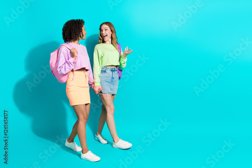 Two young women walking and chatting in trendy casual outfits against a vibrant turquoise background photo