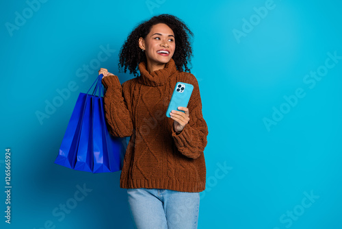 Young woman holding shopping bags and smartphone while smiling against a bright blue background photo