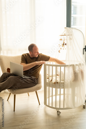 man having a home office with laptop in his lap multitasking by also taking care of the baby in the bed