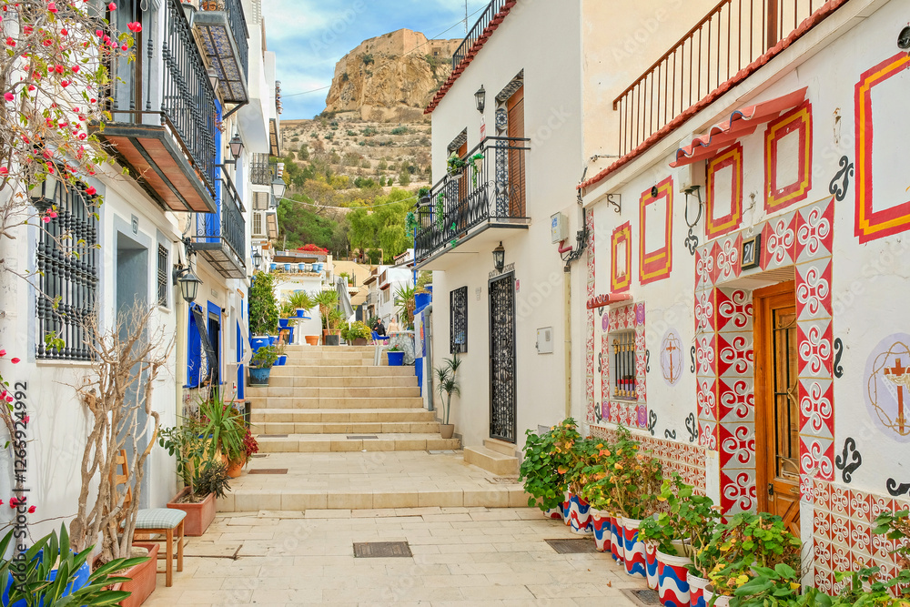 Obraz premium Narrow cozy street with steps, white houses, potted plants in historic Santa Cruz neighborhood of Alicante Old Town, located near Santa Barbara Castle