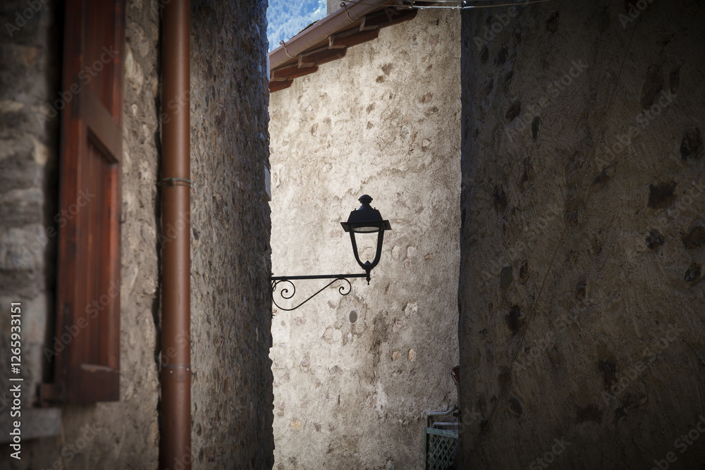 A vintage street lamp attached to a rustic stone wall of narrow old town street, featuring detailed ironwork and a weathered charm.