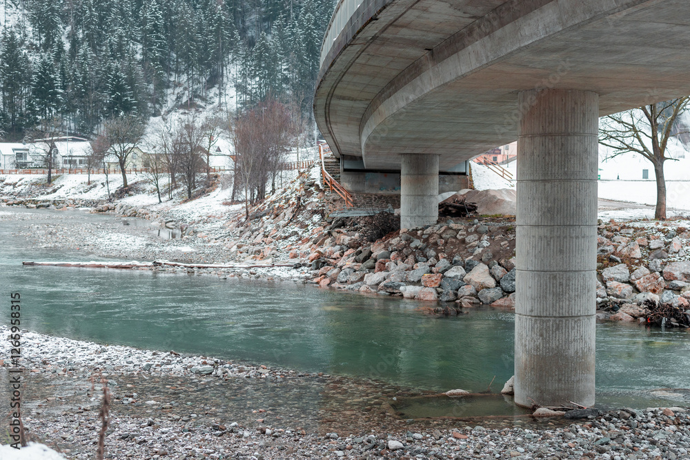 Concrete Bridge with metal railing is visible crossing the river and snow capped hills with forest seen in the background. Detail on big concrete pillar