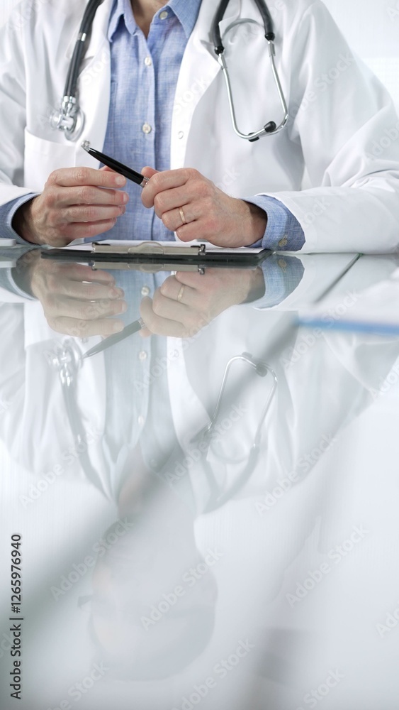 Unknown male doctor wearing a stethoscope, blue shirt and a lab coat is gesturing with hands overs a glass desk with laptop in clinic office, vertical closeup view. Medicine concept