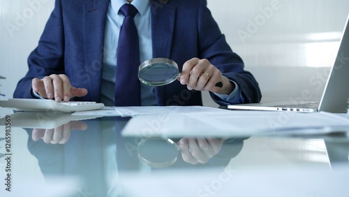 Businessman analyzing financial data with magnifying glass and calculator, conducting audit or review in office. Audit and taxes theme in business