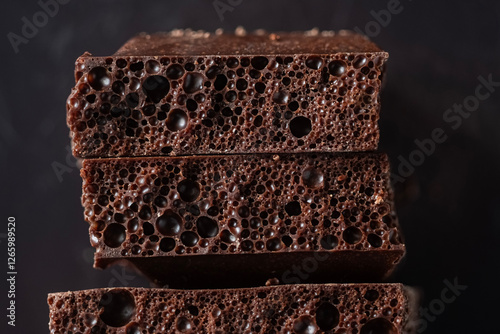 Macro of brown dark porous chocolate. Three stacked pieces against a dark background. Photo showcases the unique texture and deep color of bubbly chocolate. Soft focus. 