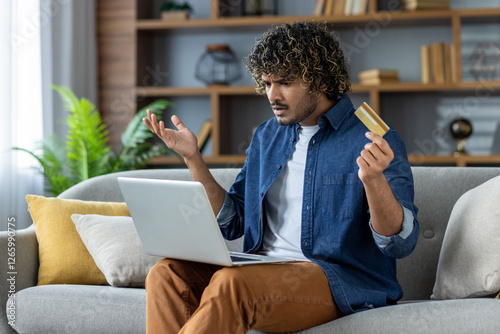 Disappointed deceived man sitting at home on sofa in living room holding bank credit card. Account blocking and fraudulent actions, failure in online money transfer.