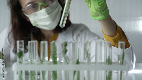 Woman scientist with dark hair wearing a lab coat, green gloves and mask holding a test tube with plants inside, close up. Science and medicine concepts